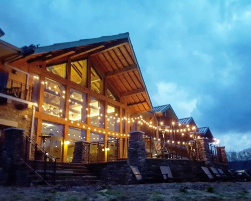 A cozy lodge with large windows and string lights, set against a cloudy evening sky.