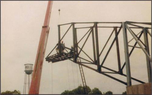 Workers on a steel framework being lifted by a crane, with a water tower visible in the background.