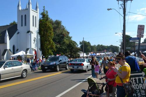 A busy street scene with a church, cars, and people enjoying a community event on a sunny day.