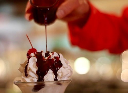 A hand pours chocolate syrup over a sundae topped with whipped cream and a cherry.
