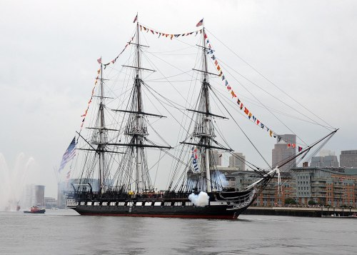 A historic tall ship sails on calm waters, adorned with colorful flags, with a city skyline in the background.