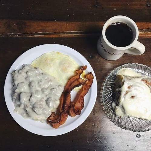 A plate with eggs, bacon, and biscuits smothered in gravy, alongside a cup of coffee and a cinnamon roll.