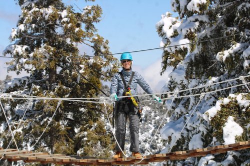 A person in a blue helmet and harness walks on a suspended wooden bridge surrounded by snow-covered trees.