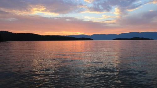 A serene lake at sunset, with colorful reflections on the water and silhouettes of distant mountains and islands.