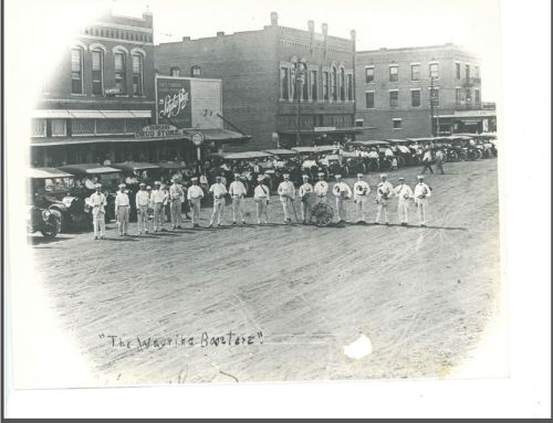 Historic black-and-white photo of a marching band in a town square, with buildings and spectators in the background.