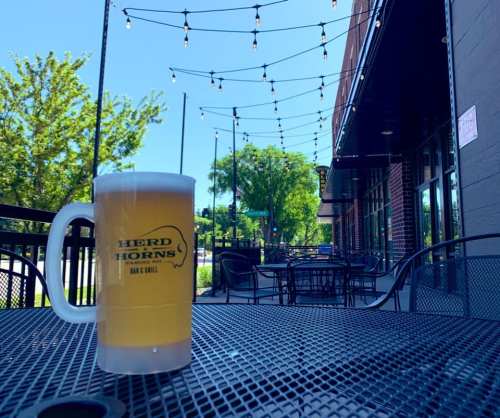 A frosty beer mug sits on a table outside a bar and grill, with string lights and greenery in the background.