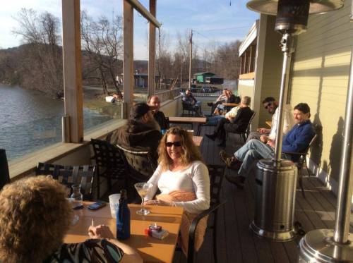 A group of people relaxes on a sunny deck by a river, enjoying drinks and conversation.