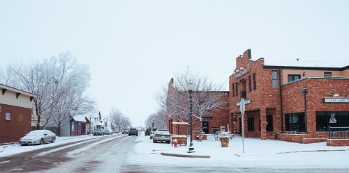 A snowy street scene featuring brick buildings, parked cars, and bare trees in a small town.