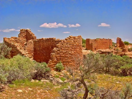 Ruins of ancient stone structures surrounded by greenery under a clear blue sky with scattered clouds.