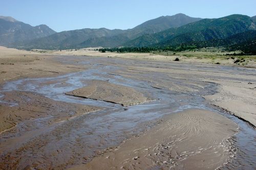 A winding stream flows through a sandy landscape, with mountains in the background under a clear blue sky.