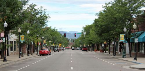 A quiet street lined with trees, shops, and traffic lights, with mountains visible in the background under a cloudy sky.