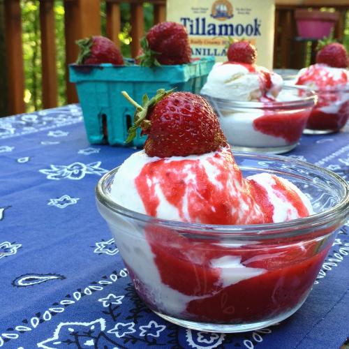 A glass bowl of vanilla ice cream topped with strawberry sauce and a fresh strawberry, set on a blue tablecloth.