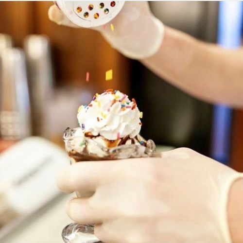 A hand in gloves adds colorful sprinkles to a sundae topped with whipped cream and chocolate sauce.
