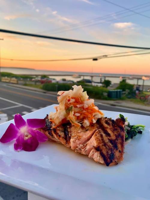 Grilled salmon topped with coleslaw, served on a white plate with a sunset view over the water in the background.