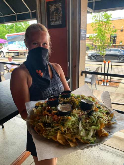 A masked server holds a large platter of nachos topped with lettuce, olives, and various sauces in a restaurant setting.