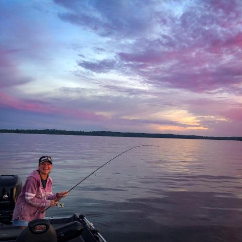 A person fishing from a boat at sunset, with vibrant pink and purple clouds reflecting on the calm water.