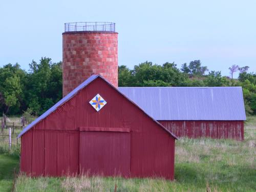 A red barn with a blue roof and a silo, surrounded by green grass and trees under a clear sky.