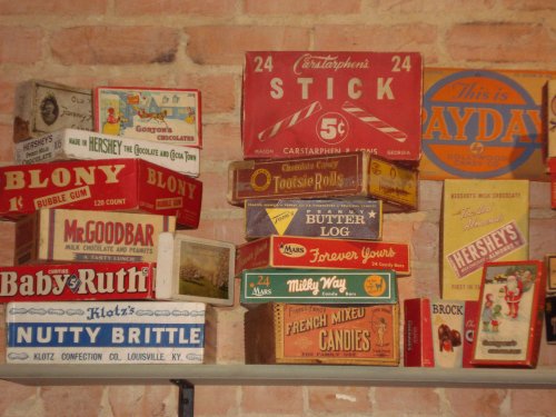 A collection of vintage candy boxes displayed against a brick wall, featuring brands like Hershey's, Baby Ruth, and Tootsie Rolls.