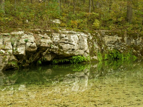 A serene riverbank with clear water reflecting rocks and lush greenery, surrounded by trees in a tranquil setting.