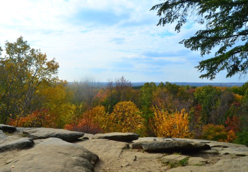 A scenic view from a rocky outcrop, showcasing vibrant autumn foliage in various shades of orange, yellow, and green.