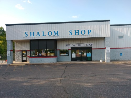 Exterior of a building with "Shalom Shop" and "Shalom Thrift Shop" signs, featuring large windows and a parking lot.
