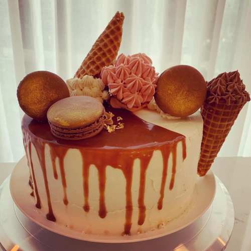 A decorated cake topped with macarons, ice cream cones, and pink frosting, featuring caramel drips.