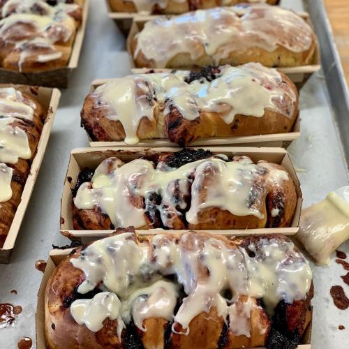 Freshly baked sweet rolls drizzled with creamy icing, displayed in wooden trays on a baking sheet.