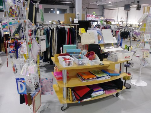 A display of various office supplies and craft materials on a cart, surrounded by clothing racks in a store.
