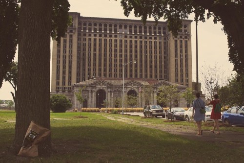 Two people walk past a large, abandoned building, framed by trees and parked cars in a grassy area.