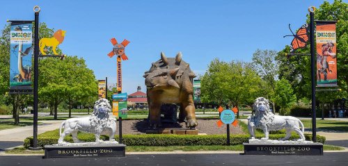 A large dinosaur statue at Brookfield Zoo, flanked by lion sculptures and colorful banners in a green park setting.