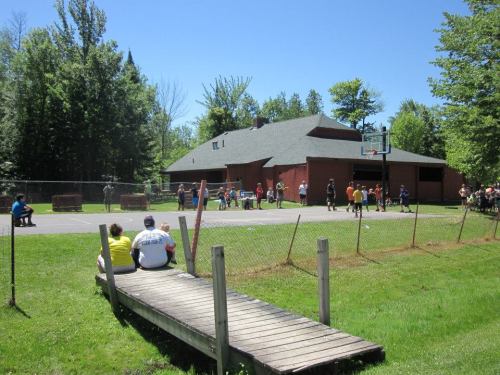 A sunny day at a park with people playing basketball and others sitting on a wooden dock nearby.