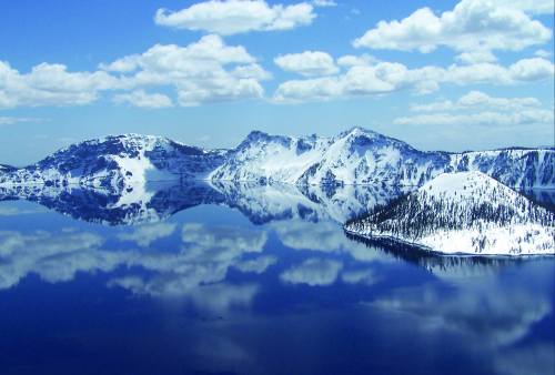 A serene landscape of snow-capped mountains reflecting in a calm blue lake under a partly cloudy sky.