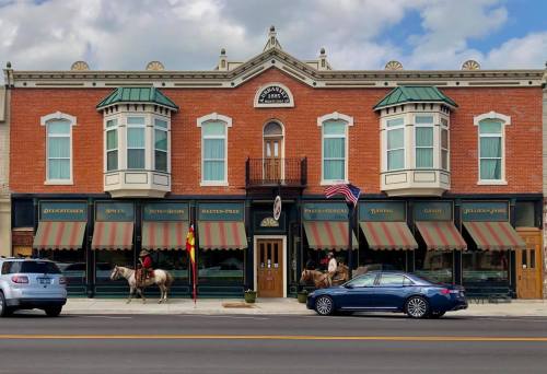Historic brick building with large windows, featuring shops and two horses tied outside, under a partly cloudy sky.