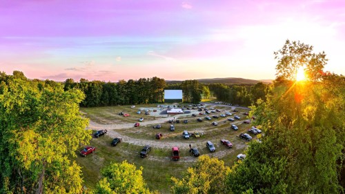 Aerial view of a drive-in movie theater at sunset, with cars parked in a circular layout and a large screen in the center.