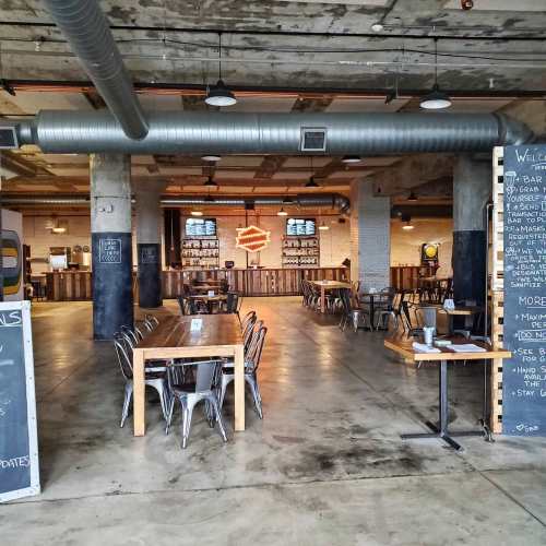 Interior of a modern restaurant with wooden tables, metal chairs, and a bar area featuring neon signage and a concrete ceiling.