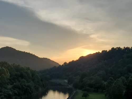 Sunset over a serene river, framed by lush green hills and a cloudy sky.