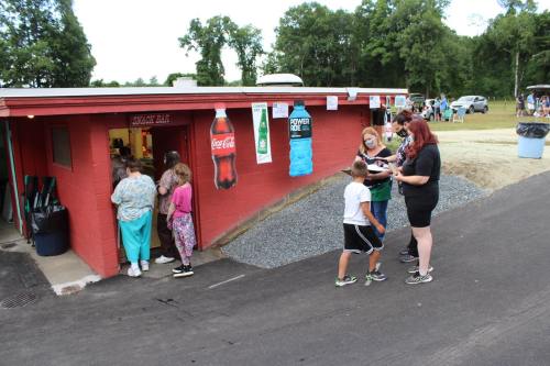 A group of people at a snack bar, with children and adults waiting in line outside a red building in a park setting.