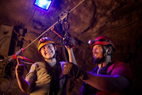 Two people in helmets and harnesses smile while preparing for a zip line in a dimly lit cave.