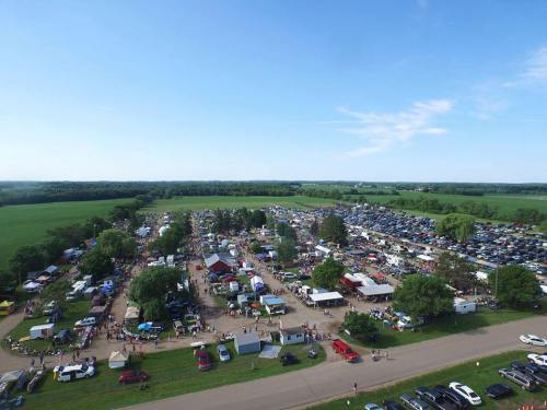 Aerial view of a bustling outdoor market with numerous tents, cars, and people amidst green fields and blue skies.