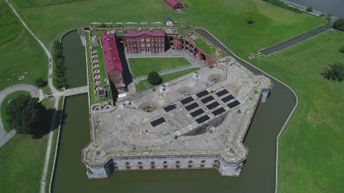 Aerial view of a historic fort surrounded by water and green fields, featuring a building with a red roof and solar panels.