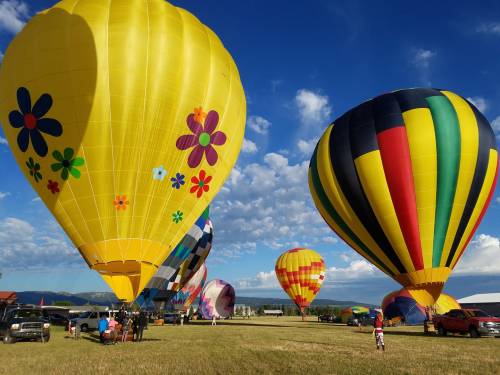 Colorful hot air balloons with floral designs and stripes fill the sky against a blue backdrop, with people and vehicles below.