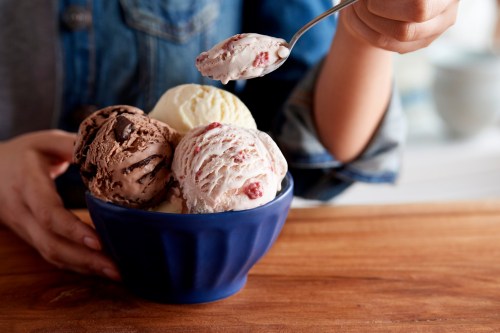 A hand holds a spoon above a bowl filled with scoops of chocolate, vanilla, and strawberry ice cream.