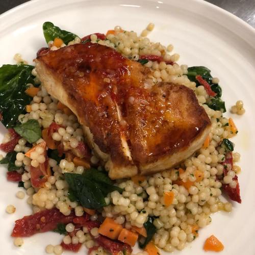 A piece of glazed fish atop a bed of couscous with spinach, sun-dried tomatoes, and diced vegetables.