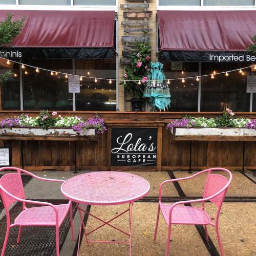 Outdoor seating area of Lola's European Cafe with pink chairs, a small table, and flower planters under string lights.