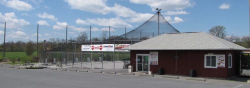 A red building with a sign, next to a large outdoor sports facility with netting and a blue sky in the background.