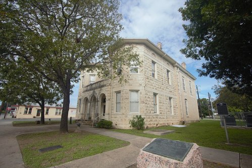 Historic stone building with arched windows, surrounded by trees and grass, under a partly cloudy sky.