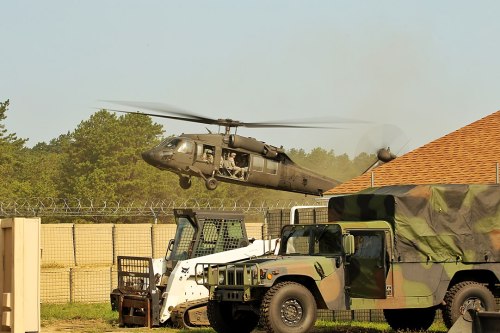 A military helicopter hovers above a fenced area, with military vehicles parked nearby. Dust is kicked up from the ground.