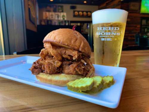 A crispy fried chicken sandwich with pickles on a plate, alongside a glass of beer in a bar setting.