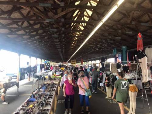 A bustling market with vendors selling clothes and accessories, featuring shoppers wearing masks in a covered area.