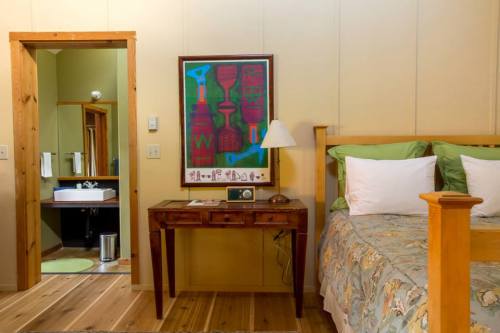 Cozy bedroom with a wooden bed, green pillows, a desk, and a bathroom doorway in the background. Colorful wall art above the desk.